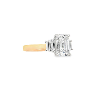 Close-up of emerald-cut lab grown diamond held by talon claws in a white gold setting, flanked by trapezoid cut side diamonds on a tapered yellow gold band, displayed on a white background.