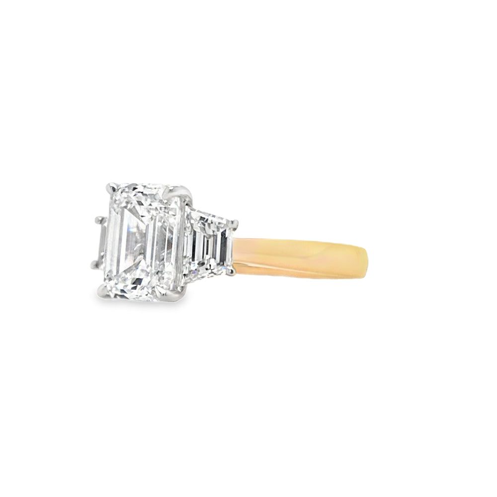Close-up of emerald-cut lab grown diamond held by talon claws in a white gold setting, flanked by trapezoid cut side diamonds on a tapered yellow gold band, displayed on a white background.
