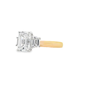 Close-up of emerald-cut lab grown diamond held by talon claws in a white gold setting, flanked by trapezoid cut side diamonds on a tapered yellow gold band, displayed on a white background.