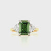 A three-stone ring featuring a central emerald-cut fancy green diamond flanked by two trillion-cut clear diamonds on a yellow gold and white gold band, set against a plain white background.
