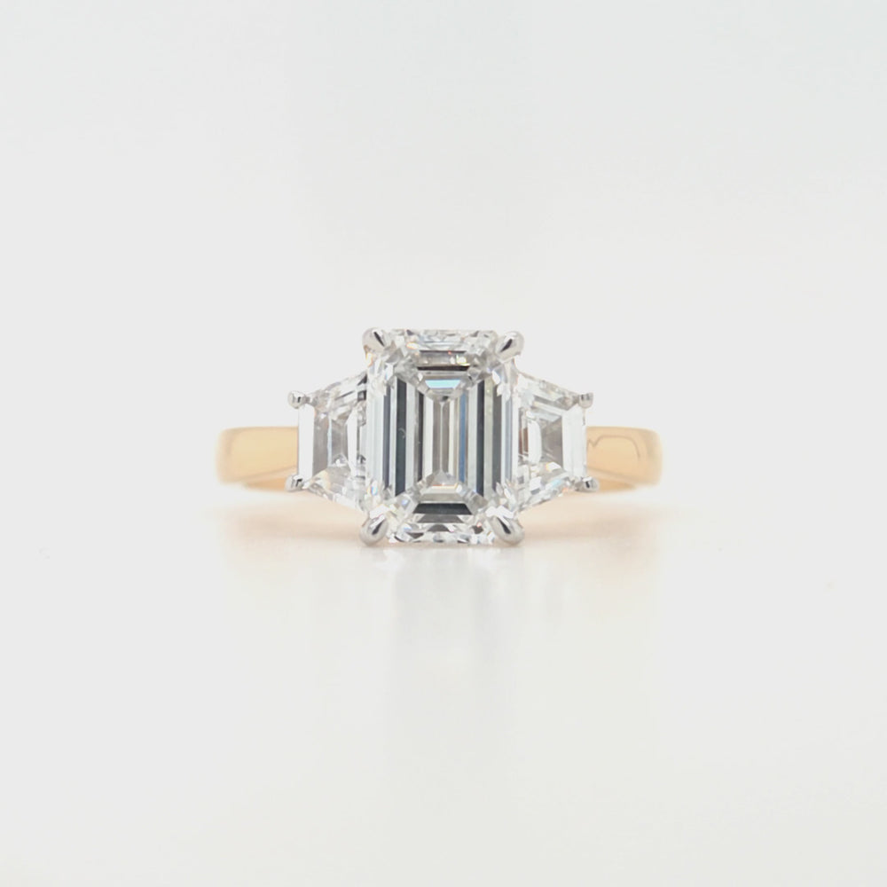 
                
                    Load and play video in Gallery viewer, Close-up of emerald-cut lab grown diamond held by talon claws in a white gold setting, flanked by trapezoid cut side diamonds on a tapered yellow gold band, displayed on a white background.
                
            