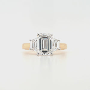 
                
                    Load and play video in Gallery viewer, Close-up of emerald-cut lab grown diamond held by talon claws in a white gold setting, flanked by trapezoid cut side diamonds on a tapered yellow gold band, displayed on a white background.
                
            