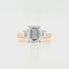 Close-up of emerald-cut lab grown diamond held by talon claws in a white gold setting, flanked by trapezoid cut side diamonds on a tapered yellow gold band, displayed on a white background.