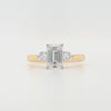 An emerald-cut and two round brilliant-cut lab grown diamonds in a platinum claw setting with a tapered yellow gold shank